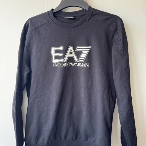 Svart sweatshirt från Emporio Armani EA7 - Säljer en sweatshirt från Emporio Armani EA7 med stort tryck på bröstet. Tröjan har rund halsringning, långa ärmar och är tillverkad i mjuk bomull.  QR-kod i tröjan garanterar dess äkthet. Fint skick. Knappt använd! 