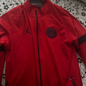 Röd PSG träningsjacka från Jordan - Säljer en röd träningsjacka från Jordan x PSG med dragkedja och svarta detaljer. Jackan har lång ärm, två fickor med dragkedja och PSG-logga på bröstet. Perfekt för fotbollsträning eller som sportig streetwear.
