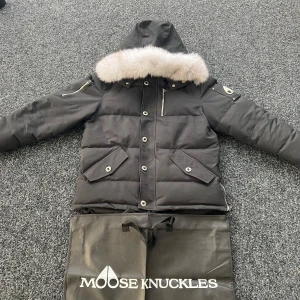 Moose Knuckles Jacka - Säljer en svart dunjacka från Moose Knuckles med avtagbar pälskrage och flera fickor med knappar och dragkedja. Jackan har lång ärm och klassisk design med logga på ärmen. Perfekt för kalla dagar. Som ny