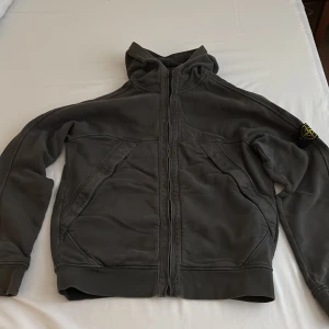 Grå hoodiejacka från Stone Island - Säljer en grå/mörkgrön hoodiejacka från Stone Island med dragkedja och huva. Jackan har den klassiska Stone Island-loggan på ärmen och stora fickor framtill. Perfekt för dig som gillar streetwear och vill ha något stilrent och bekvämt.