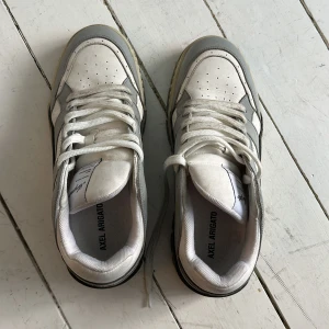 Vita och grå sneakers från Axel Arigato - Säljer ett par snygga sneakers från Axel Arigato i vitt och grått. Skorna har snörning, rund tå och en rejäl platt sula. Tillverkade i en mix av skinn och mocka med diskreta detaljer och logga på sidan. Perfekta för en stilren vardagslook. Storlek 43