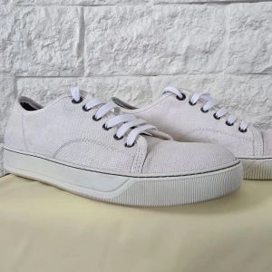 Vita sneakers från Lanvin - Säljer ett par stilrena vita sneakers från Lanvin med canvas-yttermaterial och vita snören. Skorna har rund tå och platt sula, perfekt för en clean och enkel look. Metallfärgade öljetter ger en snygg detalj. Stolek 41 men passar 42 oxå lite stora i storlikar , skorna är i  nyskick använd ett par gånger 