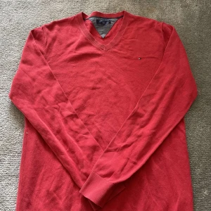 Röd v-ringad tröja från Tommy Hilfiger - Skick: 7/10 inget fel på den förutom det lilla hålet. Pris:299kr. Har du några frågor svarar vi gärna på dem