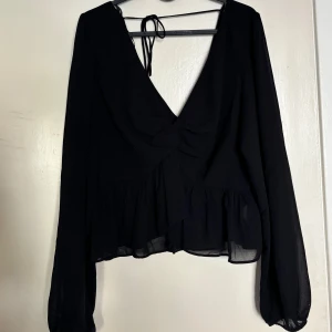 Svart blus med volang och knytband - Säljer en svart blus från H&M med v-ringning och knytband i ryggen. Blusen har långa, transparenta ärmar och volang nertill samt med en knytdetalj fram. Perfekt för dig som gillar en luftig och stilren look.                      Använd en gång, djur- och rökfritt 