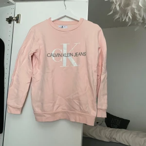 Ljusrosa sweatshirt från Calvin Klein Jeans - Säljer en ljusrosa sweatshirt från Calvin Klein Jeans med stor vit CK-logga och text framtill. Tröjan har rund halsringning och långa ärmar. Perfekt för en avslappnad och stilren look. Köpt från designonly för några år sedan för ungefär 1000kr. Knappt använd. Pris kan diskuteras vid snabb affär!!!
