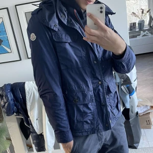 Blå vindjacka från Moncler - Säljer en blå vindjacka från Moncler med huva och flera fickor framtill. Jackan har dragkedja och knappar, samt Moncler-logga på ärmen. Perfekt för dig som vill ha en snygg och funktionell jacka med streetkänsla. Passar som S/M