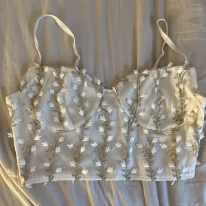 Topp  - Superfin vit croppad linnetopp med tunna axelband och broderade blommor i vitt och beige. Toppen har en lätt transparent känsla och vackra detaljer som ger en romantisk look. Perfekt för sommardagar eller festligare tillfällen.