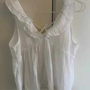 Vit volangblus från Gina Tricot - Säljer en söt vit blus från Gina Tricot med volangdetaljer runt halsen och knytning i nacken. Blusen är ärmlös och har en luftig passform, perfekt för varma dagar. Sitter som en S och aldrig använd 