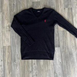 Marinblå kabelstickad tröja från Polo Ralph Lauren - Klassisk svart kabelstickad tröja från Polo Ralph Lauren med v-ringning och röd broderad logga på bröstet. Tröjan har långa ärmar och ribbade muddar vid ärmslut och nederkant. Perfekt för en stilren look.