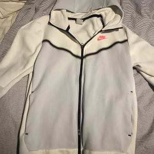 Nike tech fleece från Nike - Nike zip-hoodie i vitt och ljusgrått med svarta detaljer och dragkedja framtill. Zip-upen har huva, lång ärm och en svart dragkedjeficka på ärmen. Nike-logga i rosa på bröstet. Perfekt för träning eller chill.skick 10/10 aldrig använd,priset kan diskuteras.