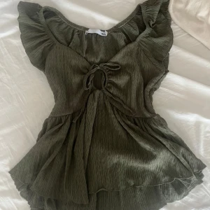 Olivgrön volangtopp med knyt - Supersöt olivgrön topp med volangärmar och volangkant nertill. Toppen har knytning framtill och en lätt rynkad struktur i tyget. Perfekt för vår och sommar!
