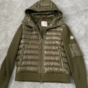 Moncler Cardigan - Snygg olivgrön cardigan från Moncler med huva och dragkedja. Storlek L men passar även storlek M. Den är i perfekt skick! Jackan har quiltad framsida och huva i glansigt material samt stickade ärmar och rygg. Ficka med dragkedja på ärmen och Moncler-logga. 