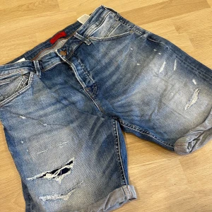 Blå jeansshorts från Jack & Jones - Snygga blå jeansshorts från Jack & Jones med slitna detaljer och upprullade benslut. Klassisk femficksmodell med knappgylf och normal passform. Perfekta för en avslappnad stil under varmare dagar.