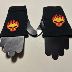 Svarta fingerlösa handskar med eldskalle onesize - Svarta fingerlösa handskar med broderad eldskalle i rött och gult ovanpå. Handskarna har en cool och edgy stil, perfekta för att sticka ut. Tunt och stretchigt material som sitter tajt runt handen. Helt nya. I stretchig bomullstrikå.