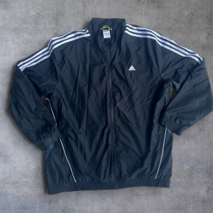 Svart vindjacka från Adidas - Säljer en svart vindjacka från Adidas med klassiska vita ränder på axlarna och dragkedja framtill. Jackan har lång ärm, ståkrage och två fickor med dragkedja. Perfekt för träning eller sportiga outfits. Svarta matchande byxor på min profil
