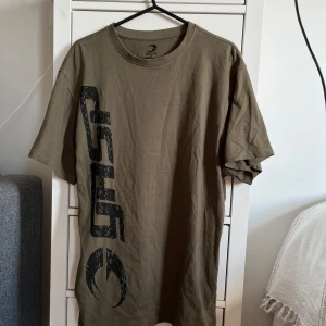 Olivgrön t-shirt med GASP-tryck - Säljer en olivgrön t-shirt från GASP med stort svart tryck både fram och bak. T-shirten har rund halsringning och är tillverkad i mjuk bomull. Perfekt för en avslappnad stil.