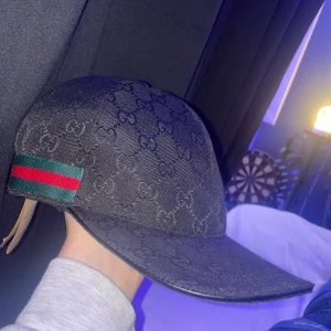 Svart keps från Gucci med monogram - Säljer en svart keps från Gucci med klassiskt GG-monogram över hela kepsen. På sidan finns ett grönt och rött band, typiskt för Gucci. Kepsen har böjd skärm och justerbar rem baktill.