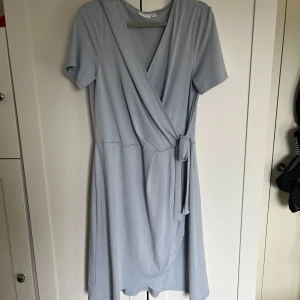 Ljusblå omlottklänning från Vila - Säljer en ljusblå omlottklänning från Vila med korta ärmar och knyt i midjan. Klänningen har en v-ringad omlottdesign och är tillverkad i ett mjukt, lätt material. Perfekt för vår och sommar!