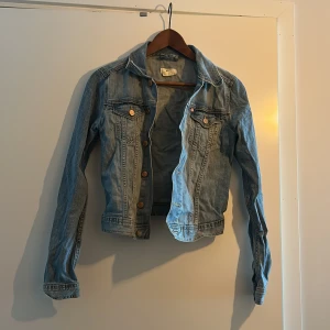Ljusblå jeansjacka från &NOW - Säljer en klassisk ljusblå jeansjacka från &NOW i damstorlek XS. Jackan har knappar framtill, två bröstfickor och långa ärmar. Tillverkad i bomull med lite stretch för extra komfort. Perfekt att slänga över en t-shirt eller klänning. 
