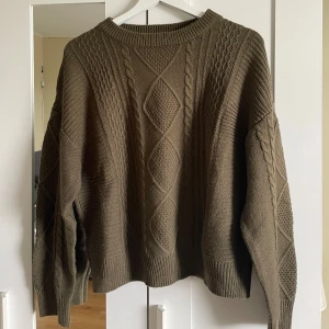 Olivgrön stickad tröja  - Säljer en olivgrön stickad tröja från DAZY med grovt kabelstickat mönster framtill och på ärmarna. Tröjan har rund halsringning, långa ärmar och ribbade muddar vid ärmslut och nederkant. Perfekt för lager-på-lager-stil.🩷