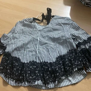 Randig blus med svart spets - Säljer en snygg blus med svartvitt randigt mönster och dekorativ svart spets längs nederkanten och ärmarna. Blusen har trekvartsärm och knytning i nacken för en extra detalj.