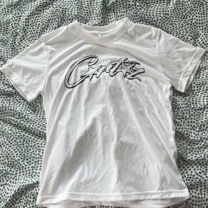 Vit t-shirt med tryck från Corteiz  - Säljer en vit t-shirt från Corteiz med ett stort svart och vitt tryck på bröstet. T-shirten har rund hals och korta ärmar, perfekt för en avslappnad stil.