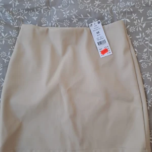 Beige kortkjol i skinnimitation XS - Snygg beige kortkjol från ginatricot i skinnimitation. Kjolen har en stilren och enkel design med raka sömmar framtill och bak. Perfekt för dig som gillar en clean och trendig look. Passar till både sneakers och boots. Lapp sitter kvar 