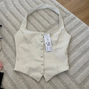 Beige omlottväst med knappar - Snygg beige väst från gina med breda axelband och markerad midja. Västen har en omlottliknande form och stängs med klädda knappar framtill. Perfekt att styla över en skjorta eller t-shirt för en trendig look.