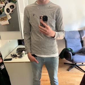 Grå långärmad tröja från Polo Ralph Lauren - Snygg tunn grå långärmad tröja från Polo Ralph Lauren med klassisk rund hals och broderad marinblå logga på bröstet. Tröjan är i bomull och har en normal passform, perfekt till jeans eller chinos för en clean och enkel stil.