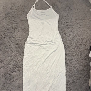Vit långklänning med smala axelband - En stilren vit långklänning med smala axelband som knyts i nacken. Klänningen har en figurnära passform och ärmlös design, perfekt för varma dagar. Materialet känns mjukt och lätt mot huden.