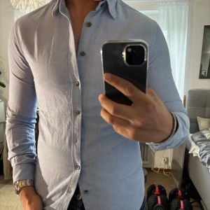 Ljusblå skjorta från Tiger of Sweden - Säljer en stilren ljusblå skjorta från Tiger of Sweden i slim fit-modell. Skjortan har klassisk krage, långa ärmar och snygga knappar framtill. Perfekt för dig som gillar en clean och modern look. Materialet är mjukt och skönt, troligtvis bomull.