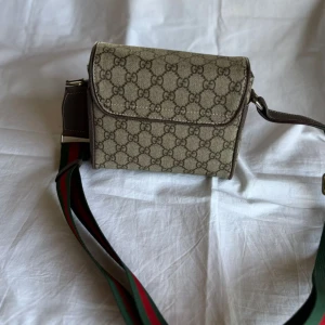 Gucci axelväska med GG-mönster - Snygg axelväska från Gucci i beige och brunt GG-mönster. Väskan har bred, justerbar axelrem i grönt och rött, klassiskt för Gucci. Ytan är i canvas med detaljer i brunt skinn och insidan är ljus. Perfekt storlek för det viktigaste.