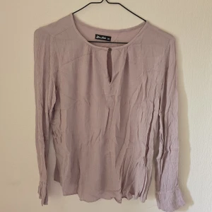 Ljuslila blus från Miss Milo XS - Supersöt ljuslila blus från Miss Milo i luftig bomull. Blusen har långärmade ärmar, rund halsringning och en liten knappdetalj framtill. Perfekt för dig som gillar en enkel men ändå stilren look.