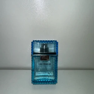 Versace Eau Fraiche parfym - Fräsch parfym från Versace, Eau Fraiche, i en stilren och modern blå glasflaska med räfflade kanter och silverdetaljer. Flaskan har en fyrkantig form och genomskinlig design som visar den ljusblå vätskan inuti.