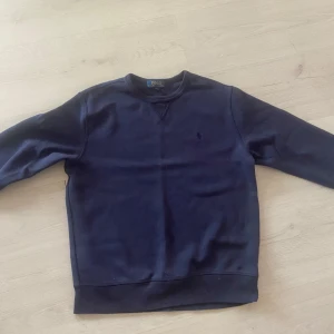 Mörkblå sweatshirt från Polo Ralph Lauren - En stilren mörkblå sweatshirt från Polo Ralph Lauren med rund halsringning och diskret broderad logga på bröstet. Tröjan har långa ärmar och är tillverkad i mjuk bomull, perfekt för en avslappnad och klassisk look.