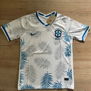 Vit Brasil Nike fotbollströja M - Snygg vit Brasilien fotbollströja från Nike med blå detaljer och grå/blå palmbladsmönster. Tröjan har V-ringning, korta ärmar och broderat CBF-märke på bröstet. Tillverkad i lätt Dri-FIT-material som andas, perfekt för match eller träning.