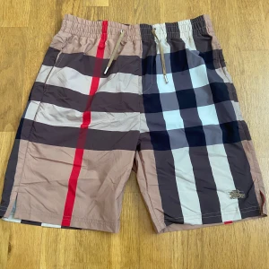 Burberry shorts/badshorts - Snygga badshorts från Burberry med klassiskt rutigt mönster i beige, svart, vitt och rött. Shortsen är nästan oanvända men jag slängde etiketten. Shortsen känns som badshorts men man kan ha på sig dem var som helst och de är väldigt lätta.