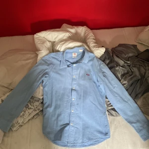 Ljusblå skjorta från Levi's, slim fit - Säljer en stilren ljusblå skjorta från Levi's i slim fit-modell. Skjortan har klassisk krage, långa ärmar och vita knappar framtill. Broderad Levi's-logga på bröstet. Perfekt för dig som gillar enkel och snygg stil.