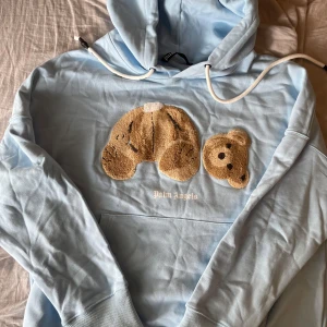 Ljusblå Palm Angels hoodie med björn - Säljer en ljusblå hoodie från Palm Angels med ikoniskt björnmotiv framtill. Tröjan har huva med vita snören, känguruficka och långa ärmar. Mjuk bomullskänsla och oversized vibe, perfekt för dig som gillar streetwear och unika detaljer. Den är I storlek M och helt oanvänd.