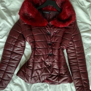 Vinröd quiltad jacka med fuskpäls - Snygg vinröd quiltad jacka från Beidifan med stor fluffig fuskpäls-krage. Jackan har markerad midja, knappar framtill och ett glansigt material som ger en lyxig känsla. 