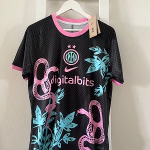 Inter Milan x Nike svart fotbollströja M - Unik Inter Milan fotbollströja från Nike i svart med rosa och turkosa detaljer. Tröjan har korta ärmar, rosa ormmotiv och turkosa blad, samt rosa kant vid hals och ärmslut. Digitalbits-logga och klubbmärke framtill. Tillverkad i lätt och ventilerande material.