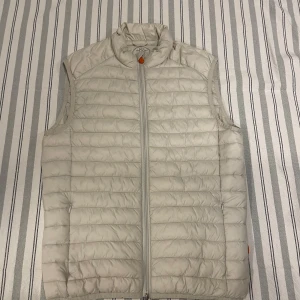 Ljusbeige dunväst från Save The Duck - Snygg ljusbeige dunväst från Save The Duck med horisontella quiltade sömmar och hög krage. Västen har dragkedja framtill och två sidofickor med dragkedja. Perfekt att slänga över en hoodie eller tröja för extra värme.
