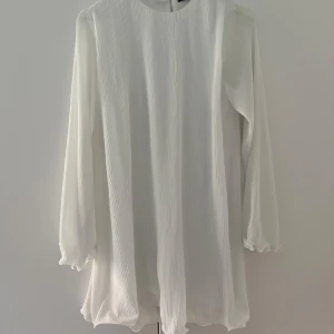 Vit plisserad klänning Gina Tricot - Superfin vit kortklänning från Gina Tricot med plisserat tyg och vida, vågiga ärmar. Klänningen har en lös passform och är tillverkad i ett lätt, flowy material som ger en luftig känsla. Perfekt för dig som gillar en stilren och trendig look. Volanger längst ner på armar och på själva klänningen. 