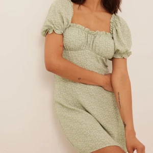 Grön blommig kortklänning från NA-KD - Supersöt ljusgrön kortklänning från NA-KD med små vita blommor, puffärmar och fyrkantig urringning. Klänningen har en lätt figursydd passform och är perfekt för vår och sommar. Tillverkad i mjuk bomull som känns skön mot huden.