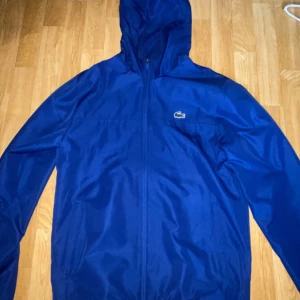  Lacoste Blå vindjacka - Snygg blå vindjacka från Lacoste Sport med huva och dragkedja framtill. Jackan har meshfoder, regular fit och klassisk Lacoste-logga på bröstet. Perfekt för dig som gillar sportig och stilren look.