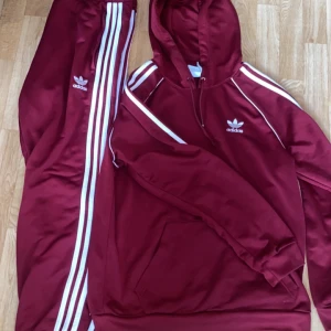 Vinröda Adidas tracksuit  - Snygga vinröda mjukisbyxor från Adidas med klassiska vita ränder längs benen och Adidas-logga på låret. Byxorna har en sportig vibe och är perfekta för chill eller träning. Matchar med hoodie för en komplett look.