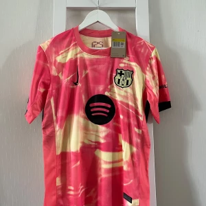 Barcelona Lamine Yamal 19 Nike fotbollströja - Snygg Barcelona fotbollströja med Lamine Yamal 19 på ryggen. Tröjan har ett unikt rosa och gulmönstrat tryck, svarta detaljer och klubbmärke på bröstet. Tillverkad i lätt och ventilerande polyester, kortärmad modell med svarta muddar och sponsorer som Spotify och Ambilight.