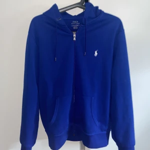 Blå hoodie från Polo Ralph Lauren - Snygg marinblå hoodie från Polo Ralph Lauren med dragkedja och huva. Klassisk vit logga broderad på bröstet, känguruficka framtill och snörning i huvan. Perfekt för en chill och stilren look.