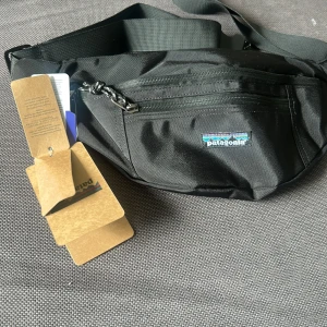 Svart magväska från Patagonia - Snygg svart magväska från Patagonia med justerbart band och dragkedja framtill. Perfekt för att bära det viktigaste och har en clean look med diskret logga på framsidan. Tillverkad i slitstark syntet och smidig att ha över axeln eller runt midjan. Helt oanvänd. 