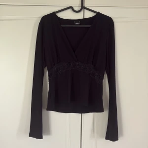 Svart omlottopp från Gina Tricot - Svart omlottopp från Gina Tricot med lång ärm och v-ringning. Toppen har en snygg spetsdetalj med blommönster under bysten som ger en extra touch. Perfekt för dig som gillar stilrena och feminina plagg.